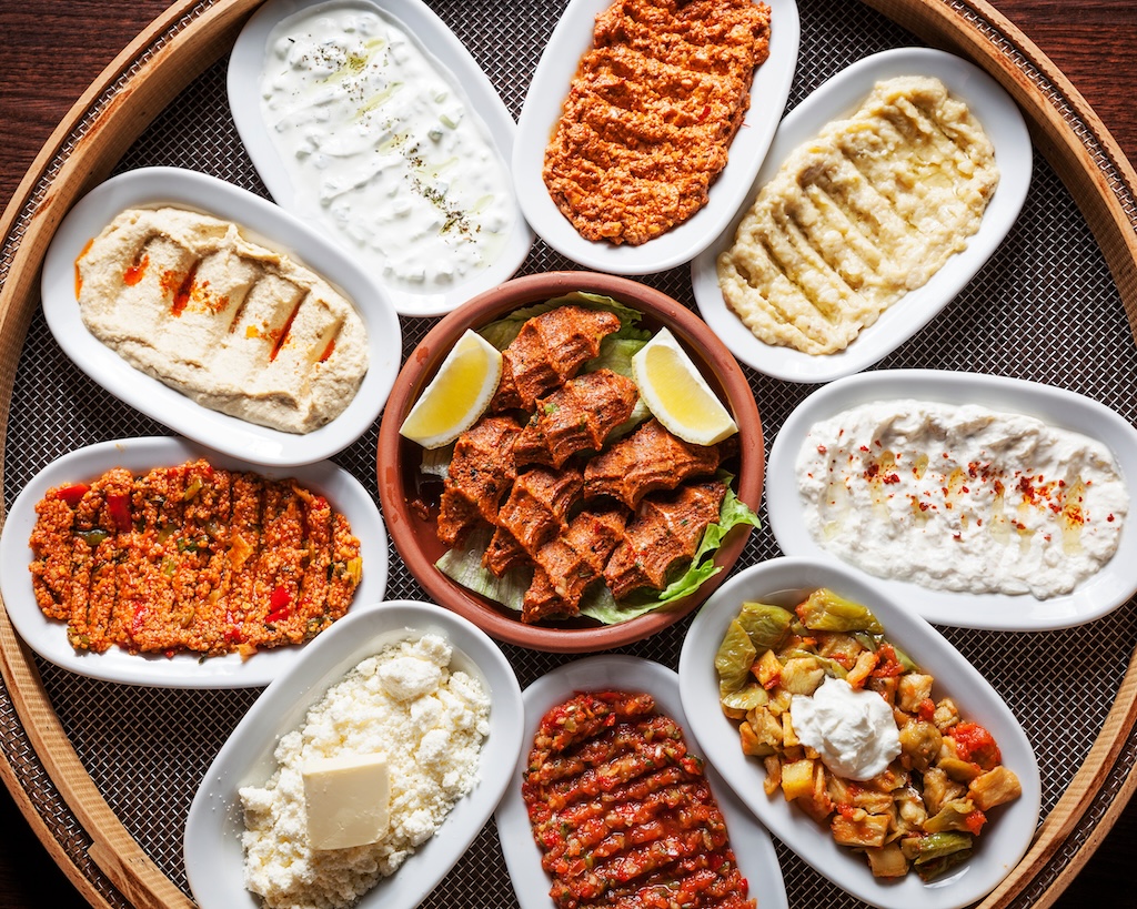 A selection of Turkish mezze including hummus, baba ganoush, spicy walnut dip, grilled meat, and more, served on a wooden table.